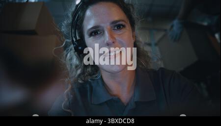 POV from Phone Screen: Female Employee Wearing Headset Using Smartphone, Scrolling and Chatting in Social Networks. In the Background, Diverse Loaders Working in Logistics Retail Warehouse at Night. Stock Photo