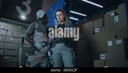 Outside of Logistics Distribution Warehouse at Night: Female Inventory Manager Using Digital Tablet, African American Employee Checking Cardboard Boxes with E-Commerce Goods in Truck for Delivery. Stock Photo