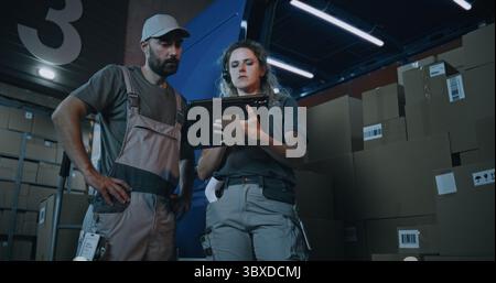Outside of Logistics Warehouse at Night: Female Inventory Specialist Using Tablet Computer, Male Loader Checking Cardboard Boxes with Online Orders, Goods in Delivery Truck for Shipping to Customers. Stock Photo