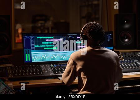 Black young adult man working at audio mixing console wearing headphones, editing music tracks on dual computer monitors in professional recording studio, focused on production Stock Photo