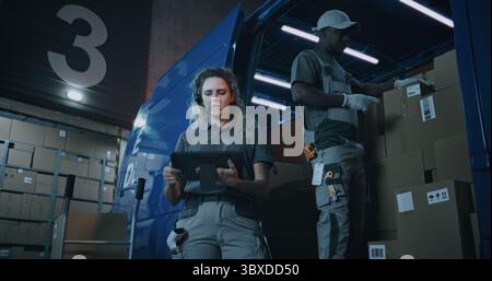 Outside of Logistics Distribution Warehouse at Night: Female Inventory Manager Using Digital Tablet, African American Employee Checking Cardboard Boxes with E-Commerce Goods in Truck for Delivery. Stock Photo