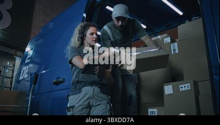 Outside of Logistics Warehouse at Night: Female Inventory Specialist Using Tablet Computer, Male Loader Checking Cardboard Boxes with Online Orders, Goods in Delivery Truck for Shipping to Customers. Stock Photo