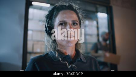 Outside of Logistics Distribution Warehouse at Night: Portrait of Female Manager Wearing Headset Looking at Camera, Working in Delivery Service. Loaders Carrying Packages in the Background. Close Up. Stock Photo