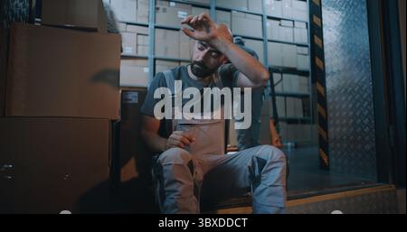 Tired Worker Carrying Cardboard Box with Online Order, Having Break, Using Smartphone. In the Background, Diverse Employees Working in Logistics Distribution Warehouse at Night. Delivery Service. Stock Photo