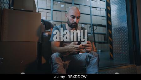 Tired Worker Carrying Cardboard Box with Online Order, Having Break, Using Smartphone. In the Background, Diverse Employees Working in Logistics Distribution Warehouse at Night. Delivery Service. Stock Photo