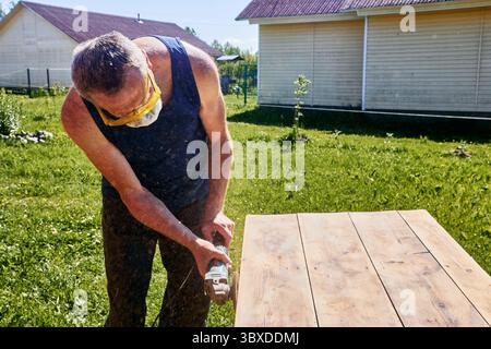 Working with handheld grinder on wooden tabletop, carpenter using protective gear during outdoor sanding in simple rural surroundings Stock Photo