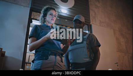 African American Employee and Female Manager Working in Express Delivery Service at Night: They Talking, Using Digital Tablet while Standing Outside of Logistics Warehouse Full of E-Commerce Goods. Stock Photo