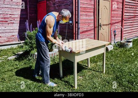 Working on tabletop edge with angle grinder, craftsman in protective gear sanding wood surface outside in rustic countryside setting Stock Photo