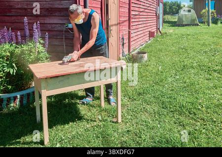 Sanding wood surface with power tool, DIYer using protective gear during backyard work in countryside setting under bright sunlight. Stock Photo