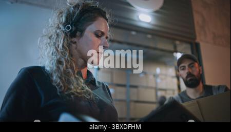 Female Manager Scanning Cardboard Boxes with Scanner, Using Tablet Computer, Working at Night. Multiethnic Workers Carrying Parcels with Online Orders, Goods from Logistics Warehouse. Slow Motion. Stock Photo