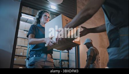 Outside of Logistics Retail Warehouse at Night: Female Worker Giving Cardboard Box with Online Orders, E-Commerce Goods Male Colleague for Delivery. Work in Express Delivery Service. Slow Motion. Stock Photo