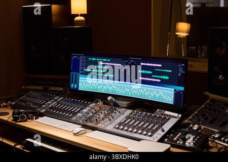Professional audio mixing console with large monitor displaying digital audio workstation interface showing multiple tracks, headphones resting on desk, studio speakers in background Stock Photo