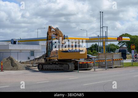 Heavy construction machinery is actively working at a busy street intersection, surrounded by barriers and piles of sand, with a fuel station in the b Stock Photo