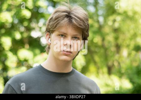 Portrait of young man standing outdoors with earbuds in public park setting, looking at camera. Summer season and green foliage as background creates Stock Photo