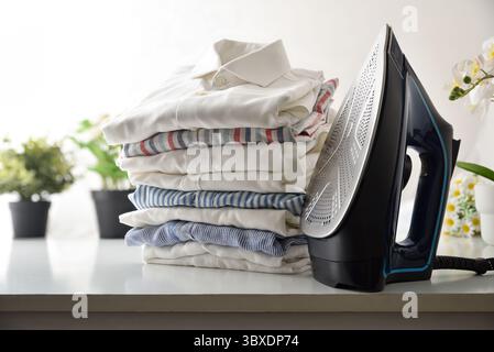 Steam iron and stack of folded shirts on a white isolated background and plants. Front view. Stock Photo