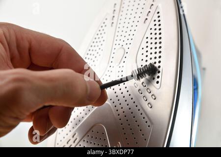 Detail of a person cleaning the steam vent holes on the metal soleplate of a steam iron with a black bristle brush on an isolated white background. Fr Stock Photo