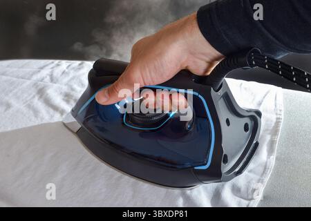 Detail of man ironing a white shirt with a steam iron on ironing board and dark isolated background. Front view. Stock Photo