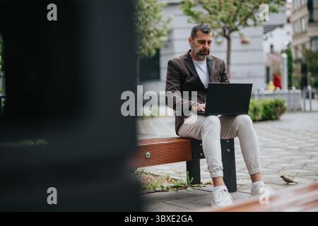 Man using a laptop on a park bench outdoors on a sunny day Stock Photo