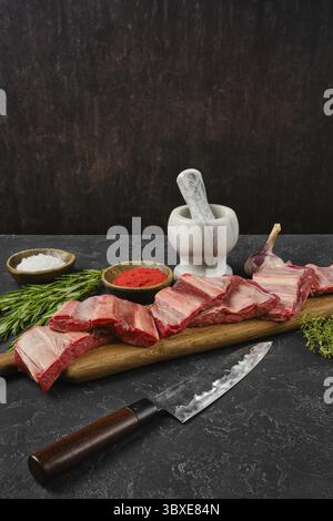 Raw beef ribs are on a wooden cutting board alongside a mortar and pestle. Herbs and spices are arranged nearby, creating an inviting atmosphere for c Stock Photo