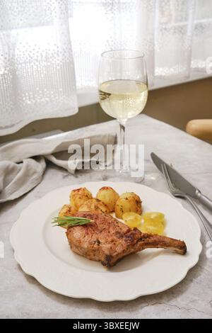 A perfectly roasted pork rib roast is plated alongside crispy golden potatoes and garnished with fresh herbs. A glass of white wine sits nearby, creat Stock Photo