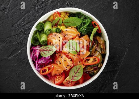 Hawaiian salmon poke bowl with rice, vegetables and seaweed seasoned with sesame seeds is served in a white bowl on a black background Stock Photo