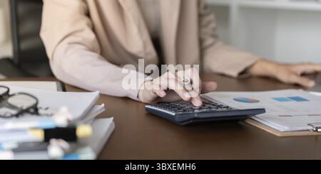A skilled accountant diligently working on financial calculations using a calculator while reviewing reports and charts on their desk in a modern offi Stock Photo