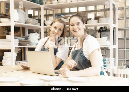 Two female entrepreneur with laptop in artisan workroom. Waist up portrait of cheerful business woman artisan looking to camera in pottery studio. Con Stock Photo
