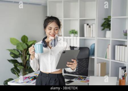 A young graphic designer in a modern office, holding a tablet and coffee, surrounded by creative tools, plants, and bookshelves Stock Photo