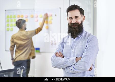 Happy smiling bearded senior developer or manager in modern IT office. Another professional working with scrum desk on background. Successful technolo Stock Photo