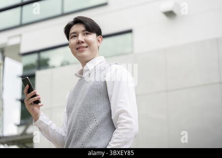 A young businessman confidently holding a smartphone while standing outside a modern office building, symbolizing success and connectivity Stock Photo