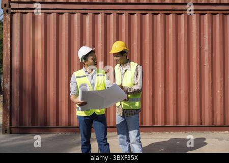 Two engineers in safety gear are reviewing plans and discussing logistics in a construction site environment, showcasing teamwork and collaboration Stock Photo