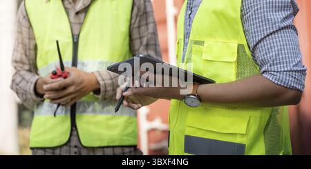 Close-up of two logistics professionals in safety vests, one holding a tablet and the other a walkie-talkie, showcasing modern technology in engineeri Stock Photo