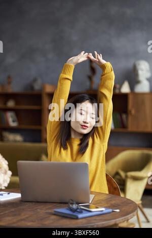 Asian woman working on laptop at home or in cafe stretching tiredly, pulling her hands up. Young lady in bright yellow jumper sits at desk. Vertical Stock Photo
