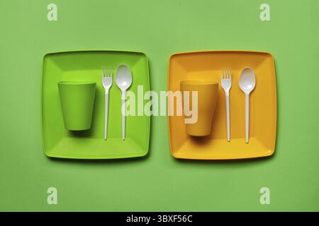 Colored plastic disposable tableware, such as two plates, spoons, forks, folded cups on green background with copy space. The concept of picnic utensi Stock Photo