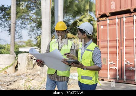 Two engineers in safety helmets and vests are smiling while reviewing logistics plans and collaborating at a shipping container site, showcasing teamw Stock Photo