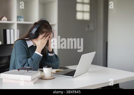 Young woman feeling stressed while wearing headphones and using a laptop in a home office Stock Photo