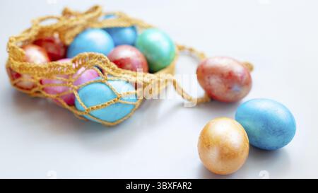 Wide banner with multi colored eggs lie in a knitted bag on white table . Three eggs lie nearby on a white background. Copy space Stock Photo