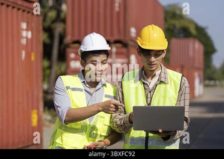Two young engineers in safety gear analyzing data on a laptop while discussing logistics strategies outdoors Stock Photo