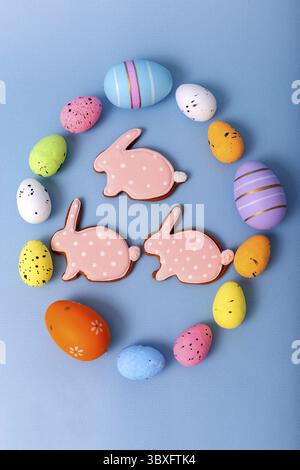 Three gingerbread in the form of a rabbit inside an oval frame of multicolored Easter eggs on a light blue background, top view Stock Photo