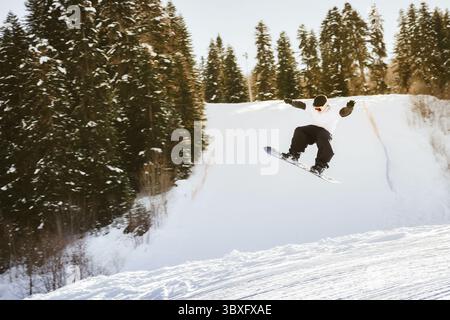 Young kid Snowboarder training in white jacket black pants jumps off freestyle on snowy slope mid-air trick forest winter freestyle ride extreme sport Stock Photo