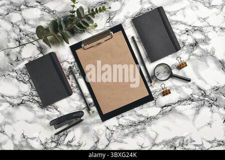 Black clipboard with brown paper, pencils, magnifier, stapler, two folders, paper clips and green twig on marble table with copy space. Education, wor Stock Photo