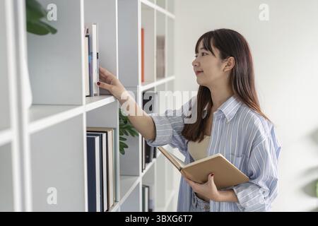 Young Student Studying Online at Home, Reaching for a Book on a Shelf, Embracing Modern Education Technology and Remote Learning Stock Photo