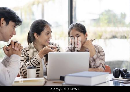 Three students working together on a laptop, sharing ideas and studying in a bright, modern environment with books and coffee Stock Photo