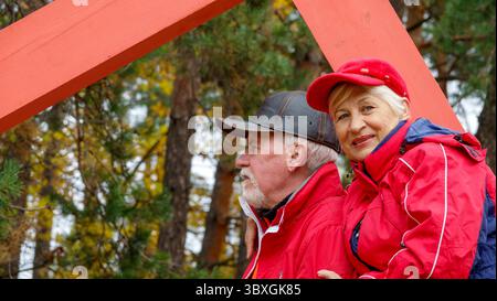 Elderly gray-haired optimistic couple in love man and woman in a landscape park in autumn in red windbreakers Stock Photo