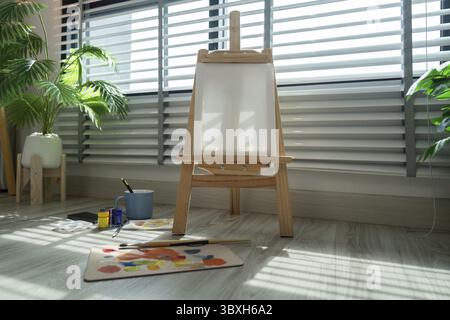 A bright artist's studio featuring a wooden easel, paints, and brushes, illuminated by sunlight streaming through large window blinds Stock Photo
