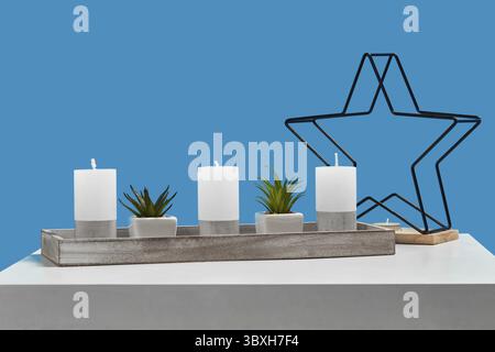 White table with green plants in pots, three big candles and one small in candlestick in form of iron star on wooden stand. Blue background. Interior Stock Photo