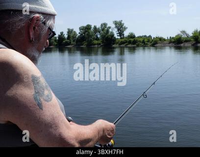 Senior man with a beanie on his head and the tattoo of a wolf on his arm is trying to catch a fish Stock Photo