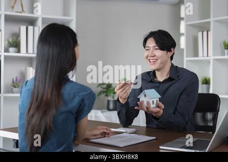 Real estate agent engaging with client, showcasing house model and keys in a contemporary office environment Stock Photo