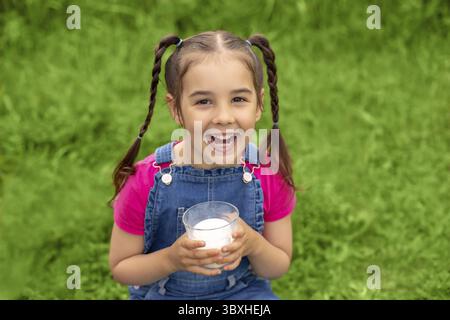 Portrait of a cute laughing brunette girl with two pigtails, in denim overalls and a pink T-shirt, on a green lawn, holds a glass with milk. Near the Stock Photo