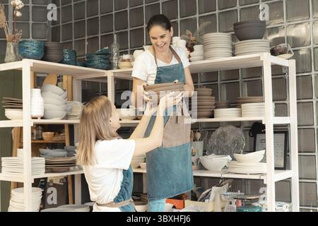 Woman with ceramic pottery. Attractive skilled young lady in apron standing at rack with dishes. Concept for workshop and master class Stock Photo